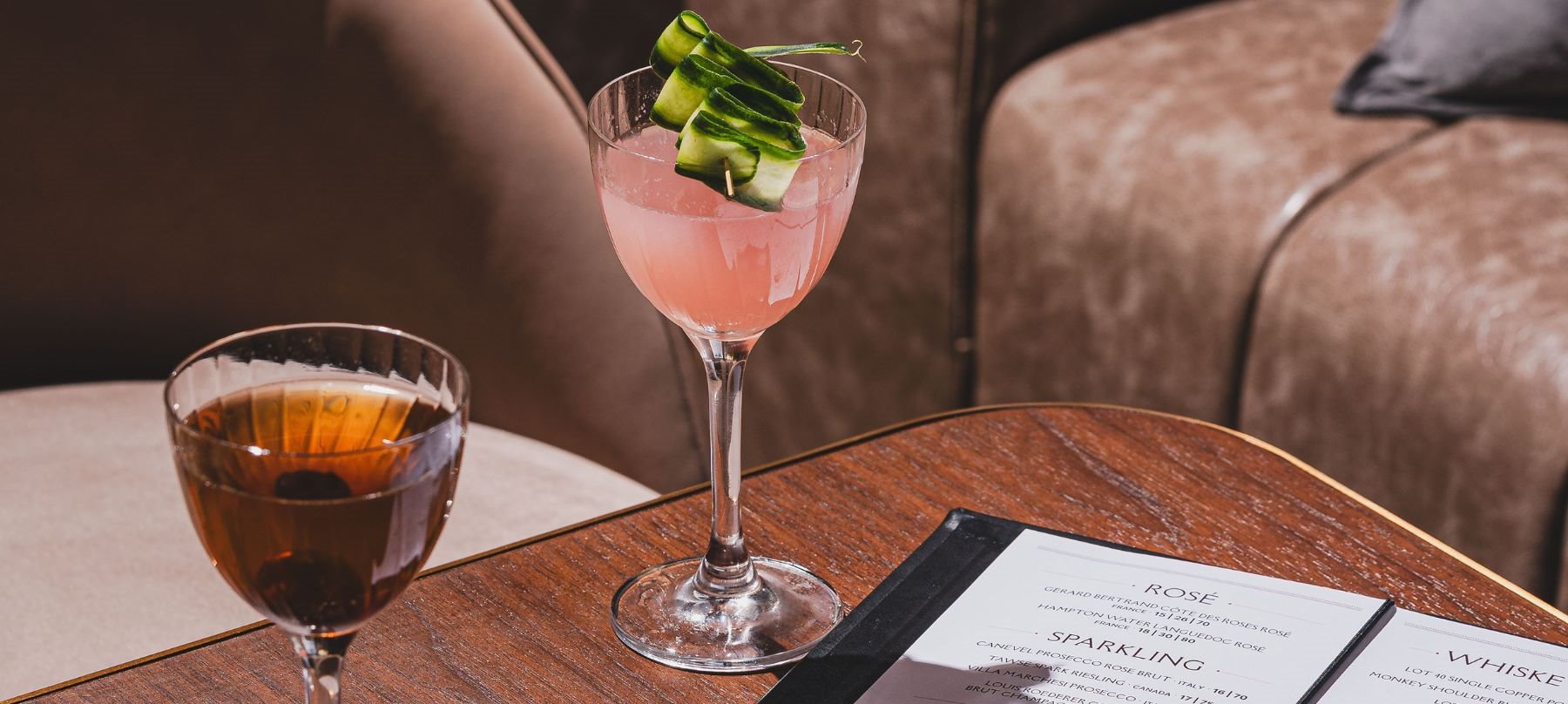 Two cocktails on a wooden table, a pink drink with cucumber garnish and a dark spirit, beside a menu in a lounge setting.